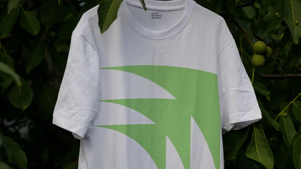 White T-shirt with a large green leaf mark, hung among dark foliage.