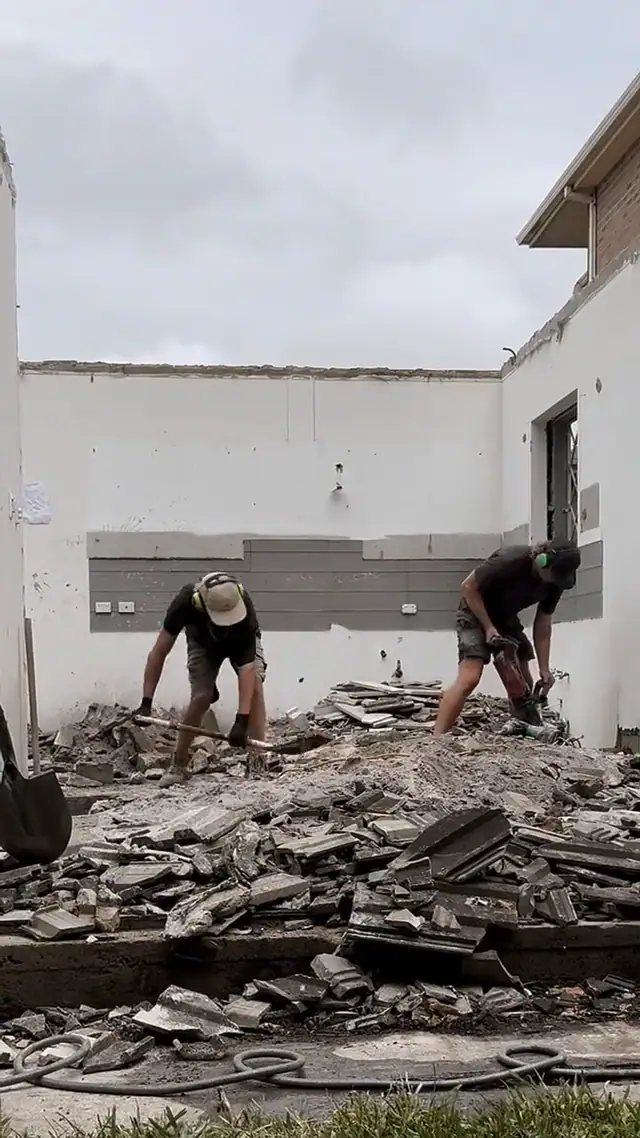 A vertical reel of tradespeople breaking up rubble and digging a post hole on a renovation site under a grey sky.